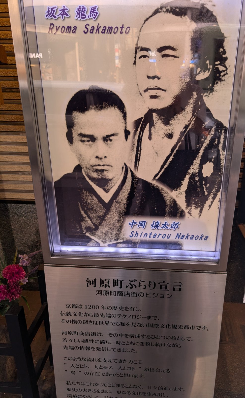 碑のそばに立つ河原町商店街振興組合の掲示。二人の肖像と「河原町ぶらり宣言」が並ぶ（2026-08-22 撮影）